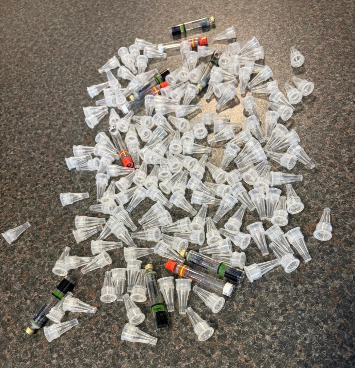 A large pile of used clear plastic insulin pen needle caps and syringes laid out on a worktop, representing one month's supply